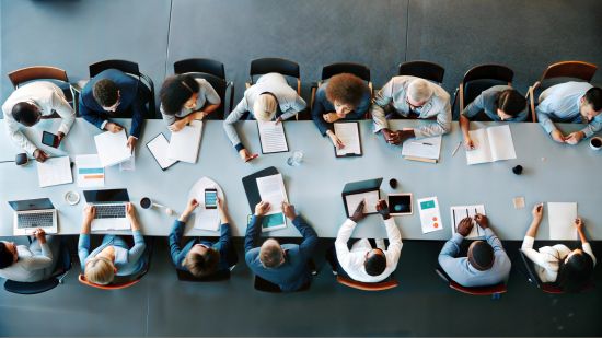 Widok z góry na długi stół konferencyjny, przy którym siedzi 12 osób w profesjonalnych strojach. Na stole rozłożone są laptopy, dokumenty, notesy, długopisy, kubki z kawą i butelki z wodą. Osoby pracują indywidualnie, przeglądając dokumenty lub korzystając z urządzeń elektronicznych. Tło stanowi jasna podłoga i minimalistyczne wnętrze biura.